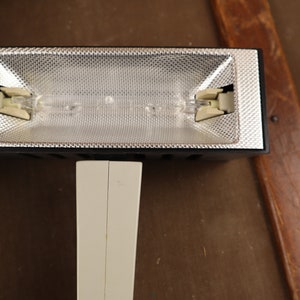 May include: A black and white studio light with a silver reflector and two bulbs. The light is mounted on a white stand.