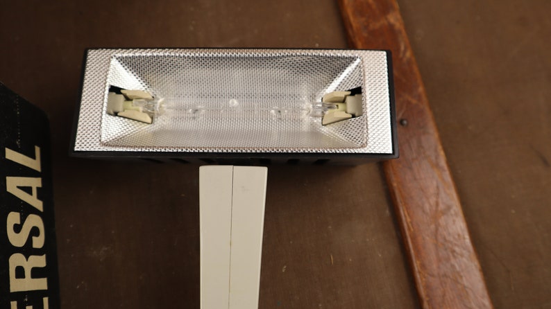 May include: A black and white studio light with a silver reflector and two bulbs. The light is mounted on a white stand.