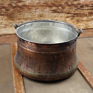 May include: A large, copper pot with two iron handles. The pot has a worn, aged appearance and is sitting on a wooden surface.