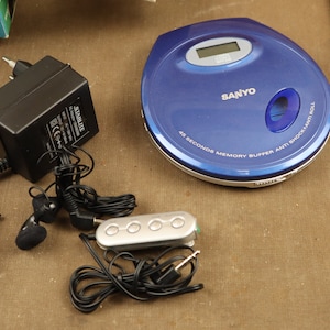 Puede incluir: Un reproductor de CD portátil azul con un adaptador de corriente negro, un mando a distancia y unos auriculares. El reproductor de CD tiene el texto "SANYO" y "45 SECONDS MEMORY BUFFER ANTI SHOCK" impreso en él.