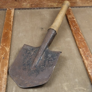 Puede incluir: Una pala de metal oxidada con un mango de madera. La pala está sobre una superficie de madera.