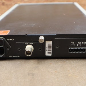 May include: Black electronic device with a power cord, a thin coax connector, and a 6-pin connector labeled "TX", "RX", and "GND". The device has a label that reads "100-230VAC".