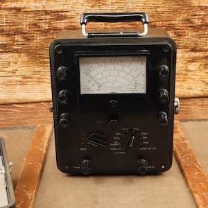 Puede incluir: Un multímetro analógico vintage negro con un dial y múltiples botones. El multímetro tiene una manija de transporte y está hecho de metal. El dispositivo está sentado sobre una superficie de madera.