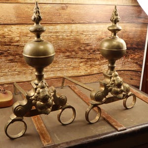 Puede incluir: Un par de fuegos de chimenea de latón antiguo con detalles ornamentados. Los fuegos tienen un remate de bola decorativo en la parte superior y un diseño de cabeza de león en la base.