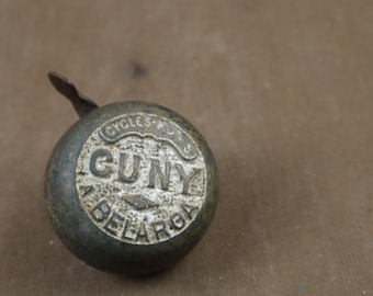 May include: A vintage bicycle bell with a silver-coloured face and the words "Guny Cycles & Bell Works" embossed on it. The bell is made of metal and has a brass-coloured lever.