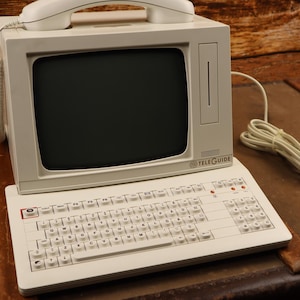 Op de afbeelding: Een witte vintage computer met een ingebouwde monitor en een toetsenbord. De computer heeft een ingebouwde telefoon en de tekst "TELEGUIDE" is zichtbaar aan de voorkant.