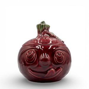 May include: A whimsical, red ceramic jar with a smiling face design. The jar has a lid with a green stem detail. The jar's face features large, circular eyes and a wide, smiling mouth. The overall design is reminiscent of a cartoon character.