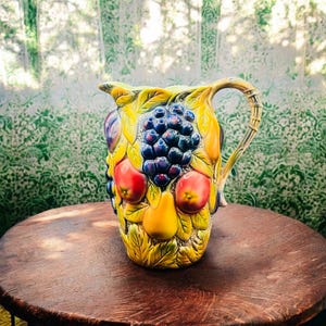 May include: A yellow ceramic jug with a handle, decorated with a colourful fruit design. The jug features pears, apples, grapes, and leaves.