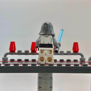 May include: A white Lego minifigure with a gray hooded robe and a blue lightsaber. The minifigure is standing on a gray platform with red lights on either side.
