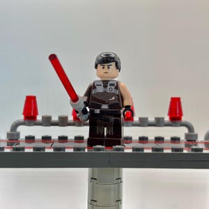 May include: A Lego minifigure of a Star Wars character with black hair, a brown and black outfit, and a red lightsaber. The minifigure is standing on a gray platform with red lights on either side.