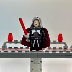 May include: A Lego minifigure of a dark-haired character wearing a black and white robe with a red cape and holding a red lightsaber. The minifigure is standing on a gray platform.