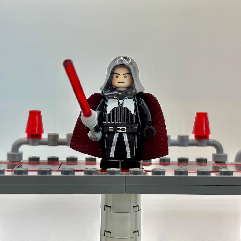 May include: A Lego minifigure of a dark-haired character wearing a black and white robe with a red cape and holding a red lightsaber. The minifigure is standing on a gray platform.