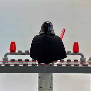 May include: A black Lego minifigure of Darth Vader standing on a gray Lego platform. The minifigure is wearing a black cape and a black helmet. The platform has red lights on either side.