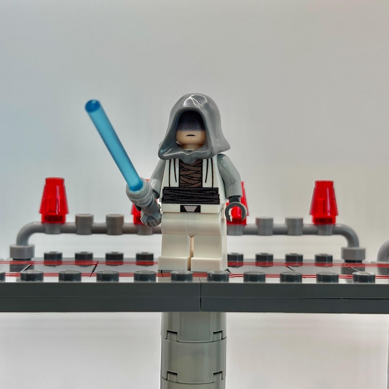 May include: A gray and white Lego minifigure with a gray hooded robe and a blue lightsaber. The minifigure is standing on a gray platform with red lights on either side.