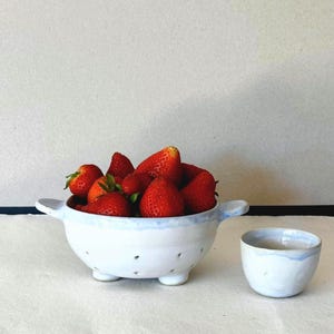 Peut inclure: Un égouttoir en céramique blanche rempli de fraises rouges fraîches. L'égouttoir a un bord bleu clair et de petits pieds. Un petit bol assorti est à côté, également avec un bord bleu. Les fraises sont mûres et prêtes à être mangées.