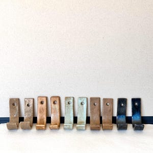 Könnte beinhalten: Zehn Keramikhaken in verschiedenen Brauntönen, Blau- und Grüntönen. Die Haken sind in einer Reihe auf einem hellbraunen Hintergrund angeordnet.