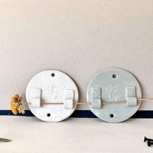 Pode incluir: Dois porta-incensos de cerâmica para montar na parede com um design floral. Os suportes são redondos e têm um furo no centro para pendurar. Um suporte é branco e o outro é cinza claro.