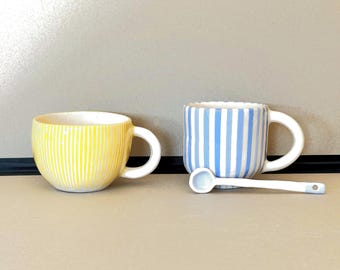 May include: Two ceramic mugs with striped patterns. One mug is yellow with white stripes and the other is blue with white stripes. A small white spoon is next to the blue mug.
