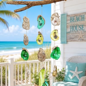 May include: A hanging mobile featuring a horizontal piece of driftwood with three vertical strands of oyster shells. Some shells are natural tan and gray, while others are painted in vibrant shades of emerald green, lime green, and yellow.