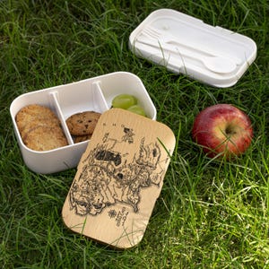 May include: White rectangular lunch box with a wooden lid featuring a map design. The box is open, revealing cookies and grapes. A red apple and a separate compartment with a fork and spoon are also visible, all set on green grass.