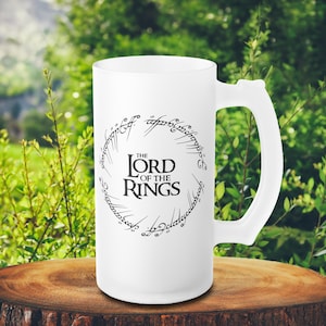 Puede incluir: Taza de cerveza blanca esmerilada con un asa grande. La taza presenta el texto "THE LORD OF THE RINGS" en negro, con una fuente estilizada, rodeado por un anillo de escritura. La taza está sobre una superficie de madera.