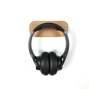 Peut inclure: Un support mural en bois pour casque audio avec un coussin en cuir brun foncé. Un casque audio noir supra-auriculaire est accroché au support.