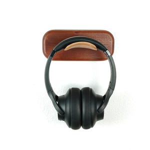 Peut inclure: Un support pour casque en bois avec une finition brune et un coussin en cuir. Un casque noir est suspendu au support.