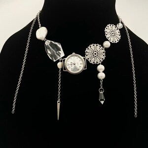 Puede incluir: Un collar plateado con un reloj, perlas y detalles de cristal. El collar tiene una cadena plateada con un reloj, dos dijes de filigrana plateada y un colgante de cristal. El collar también tiene varias perlas blancas.