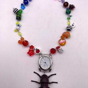 May include: A colorful beaded necklace with a variety of charms. The necklace features a silver watch pendant with the word "Geneva" on the face, and a dark brown beetle charm. Other charms include stars, dice, and fruit.