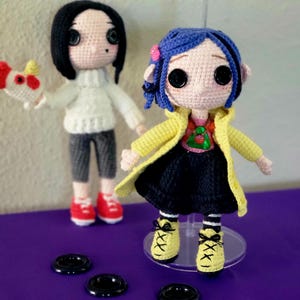 May include: Two handmade crochet dolls. One has black hair, a white jumper, grey trousers, and red shoes, holding a red and white object. The other doll has blue hair, a yellow coat, a black dress, and yellow shoes. Both dolls have button eyes.