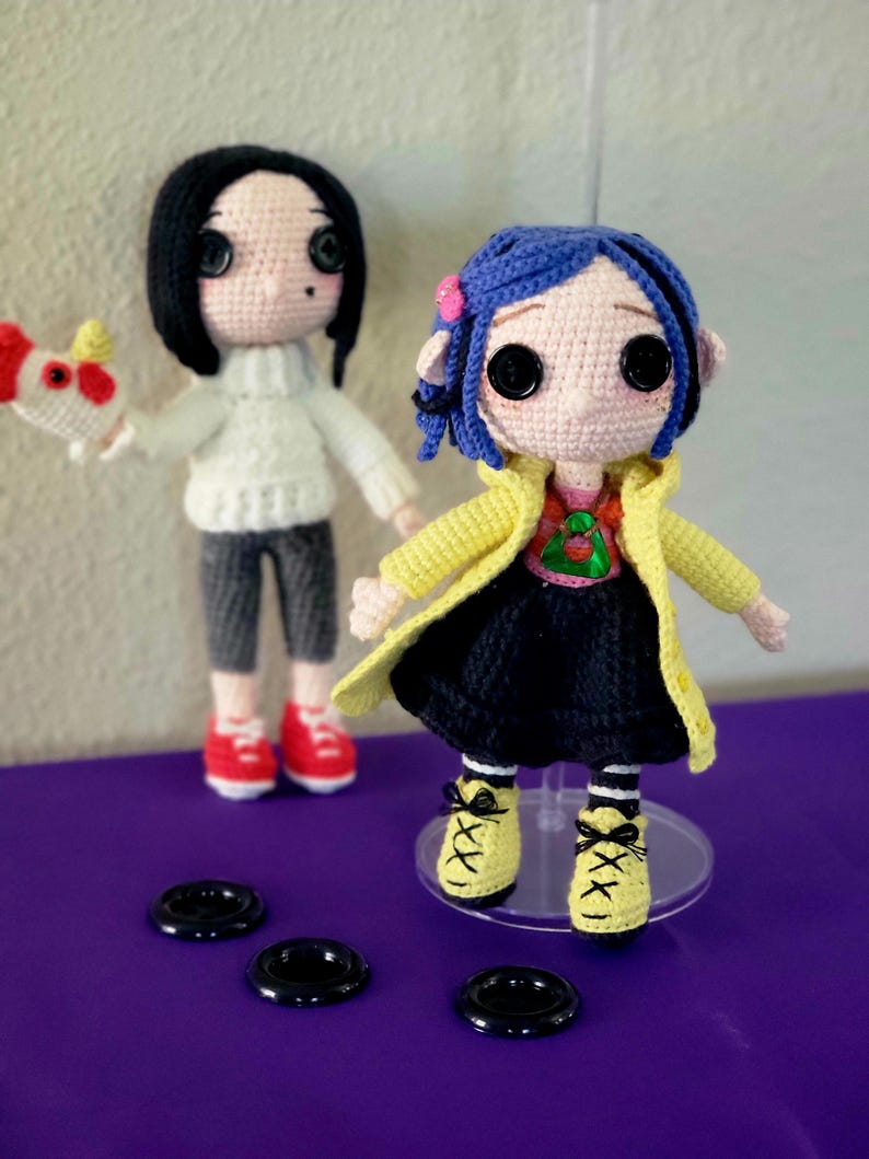 May include: Two handmade crochet dolls. One has black hair, a white jumper, grey trousers, and red shoes, holding a red and white object. The other doll has blue hair, a yellow coat, a black dress, and yellow shoes. Both dolls have button eyes.