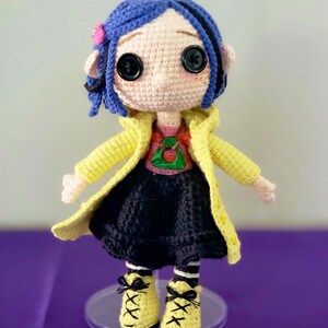 May include: A handmade crochet doll with blue hair, large black button eyes, and a pink flower. The doll wears a yellow coat, a red and green shirt, a black skirt, and yellow shoes with black laces. The doll is standing on a clear stand.
