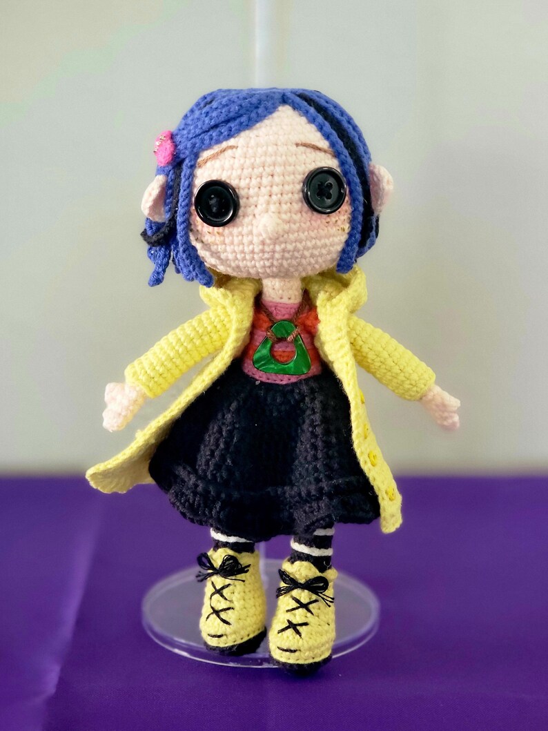 May include: A handmade crochet doll with blue hair, large black button eyes, and a pink flower. The doll wears a yellow coat, a red and green shirt, a black skirt, and yellow shoes with black laces. The doll is standing on a clear stand.
