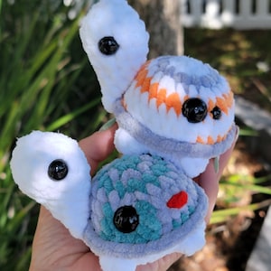May include: Three handmade plush turtles in a hand. The turtles have white heads and limbs, with colorful shells in shades of gray, teal, and orange. Each turtle has a large black eye and a small red accent.