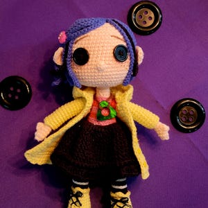 May include: A crocheted doll with blue hair, large button eyes, and a yellow coat. The doll is wearing a pink top, black skirt, and yellow shoes. Three black buttons are placed around the doll on a purple background.