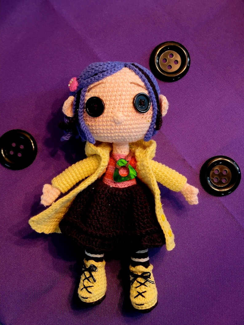 May include: A crocheted doll with blue hair, large button eyes, and a yellow coat. The doll is wearing a pink top, black skirt, and yellow shoes. Three black buttons are placed around the doll on a purple background.