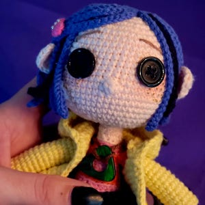 May include: Handmade crochet doll featuring button eyes, blue hair, and a yellow jacket. The doll wears a pink flower hair accessory and a red and pink shirt. The design is unique and whimsical.
