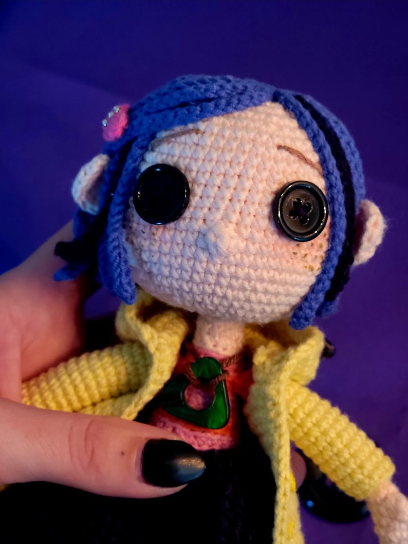 May include: Handmade crochet doll featuring button eyes, blue hair, and a yellow jacket. The doll wears a pink flower hair accessory and a red and pink shirt. The design is unique and whimsical.