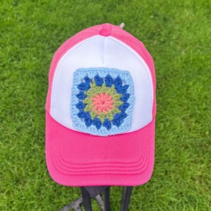 May include: A bright pink and white trucker hat featuring a square crochet patch on the front panel. The patch displays a multi-colored floral pattern in shades of light blue, dark blue, olive green, and coral pink, centered on a white background.