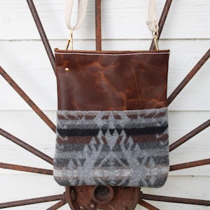 May include: A rectangular crossbody bag featuring a smooth dark brown leather upper section and a textured gray wool lower section with a geometric pattern in black, white, and gray. The bag includes a cream-colored fabric strap with gold hardware.