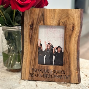 May include: A rectangular live-edge wooden picture frame featuring a natural wood grain and dark bark edges. The frame displays a photograph of three graduates in caps and gowns throwing confetti, with the text "DREAMS AND DEDICATION ARE A POWERFUL COMBINATION" printed in black at the bottom.