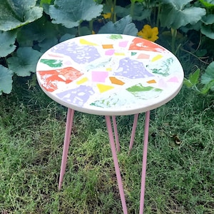 May include: A small, round, white table with pink legs. The table top is decorated with a colorful mosaic pattern of green, yellow, orange, pink, and purple.