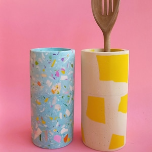 以下が含まれることがあります： ピンクの背景に、2つのセラミック製の食器入れが置かれています。1つは白とカラフルな斑点のある青色、もう1つは黄色い幾何学模様のある白色です。黄色の食器入れには木製のスプーンが入っています。
