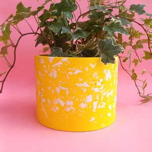 May include: A vibrant yellow cylindrical planter with a terrazzo design, filled with green and white variegated ivy. The planter is set against a pink background, creating a striking contrast. A decorative item for home decor.