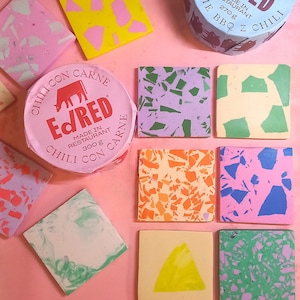 Può includere: Un set di nove sottobicchieri quadrati colorati con un design a terrazzo. I sottobicchieri sono realizzati con uno sfondo rosa chiaro, giallo chiaro, blu chiaro, verde chiaro e viola chiaro con macchie di vari colori. I sottobicchieri sono disposti in una griglia su una superficie rosa. Nell'immagine sono presenti anche due lattine di chili con carne. Le lattine sono etichettate con "Ed/Red" e "Chili con Carne".