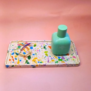 May include: A rectangular tray with a terrazzo design in white, blue, yellow, green, and orange. A light green perfume bottle and a seashell bracelet rest on the tray. The perfume bottle has the text "GO FRUITY".
