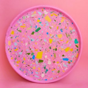 Könnte beinhalten: Ein rundes rosa Tablett mit einem Terrazzo-Design. Das Tablett ist aus Harz gefertigt und hat eine gesprenkelte Oberfläche mit blauen, grünen, gelben und weißen Flecken.