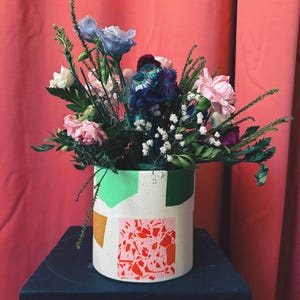 Könnte beinhalten: Ein Strauß aus rosa, blauen und weißen Blumen in einer weißen Keramikvase mit grünen, orangen und rosa geometrischen Mustern. Die Vase steht auf einem schwarzen Tisch.