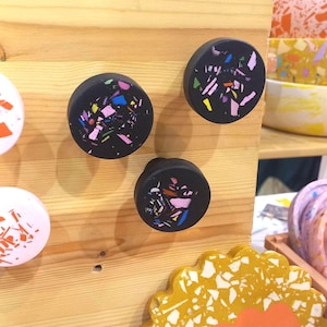 Può includere: Pomelli neri per mobili con colorati accenti in stile terrazzo montati su una superficie in legno chiaro. I pomelli hanno una forma rotonda e sono decorati con frammenti rosa, gialli, blu e verdi.