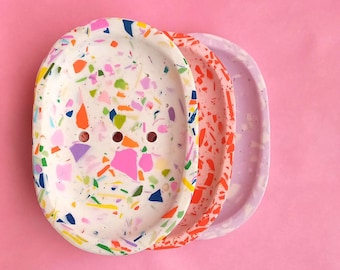May include: Three oval-shaped soap dishes stacked together. The top dish is white with multicolored terrazzo flecks in pink, orange, blue, and green. The middle dish is orange with white flecks, and the bottom dish is a solid light purple.